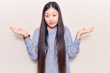 Young beautiful chinese woman wearing casual striped shirt clueless and confused with open arms, no...