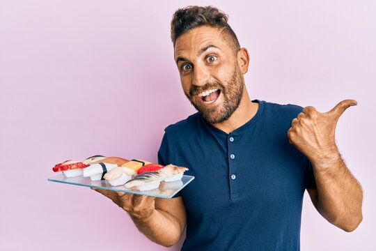 Handsome man with beard showing a plate of sushi pointing thumb up to the side smiling happy with open mouth