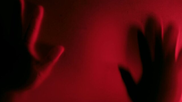 Defocused Possessed man scratching glass with hands with red light background, creepy male silhouette, horror. Scary ghostly creature behind the glass, Hand hits the matte glass surface.