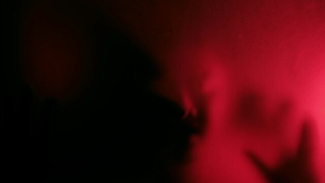 Defocused Possessed man scratching glass with hands with red light background, creepy male silhouette, horror. Scary ghostly creature behind the glass, Hand hits the matte glass surface.