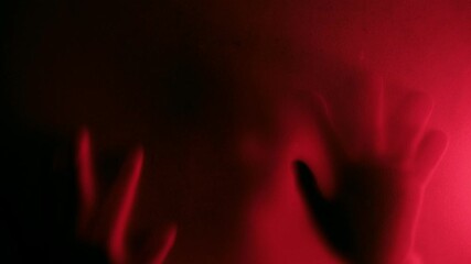 Defocused Possessed person scratching glass with hands with red light background, creepy silhouette, horror. Scary ghostly creature behind the glass, Hand hits the matte glass surface