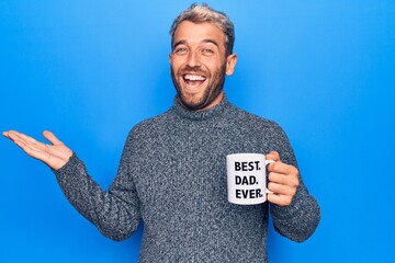 Handsome blond man drinking cup of coffee with best dad ever message over blue background celebrating achievement with happy smile and winner expression with raised hand