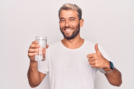 Handsome blond man with beard drinking glass of water to refreshment over white background smiling happy and positive, thumb up doing excellent and approval sign