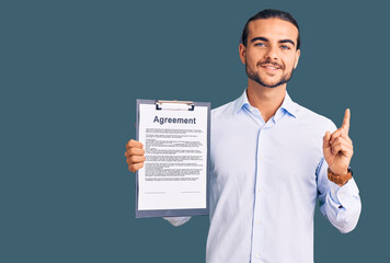 Young handsome man holding clipboard with agreement document surprised with an idea or question pointing finger with happy face, number one