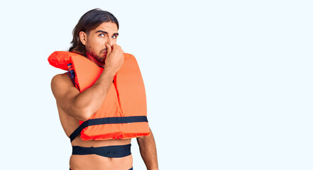 Young handsome man wearing nautical lifejacket smelling something stinky and disgusting,...