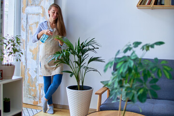 Cute happy young smiling attractive woman gardener in apron watering houseplants using spray bottle