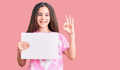 Fototapeta premium Cute hispanic child girl holding blank empty banner doing ok sign with fingers, smiling friendly gesturing excellent symbol