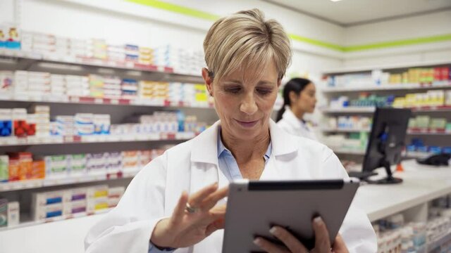 Female pharmacist adjusting settings on digital tablet while assistant sorts out scripts behind counter in pharmacy 