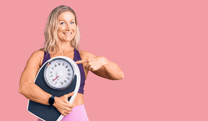 Middle age fit blonde woman wearing sports clothes holding weighing machine smiling happy pointing with hand and finger