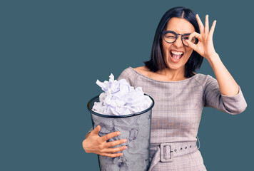 Young beautiful latin woman holding paper bin full of crumpled papers smiling happy doing ok sign with hand on eye looking through fingers