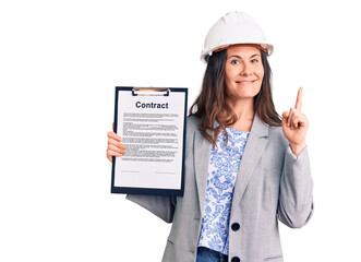 Young beautiful brunette woman wearing architect hardhat holding contract surprised with an idea or question pointing finger with happy face, number one