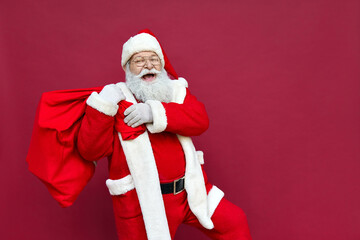 Happy old funny Santa Claus, Saint Nicholas wearing costume holding sack bag with Merry Christmas presents walking delivering xmas gifts standing isolated on red background. Xmas delivery concept.