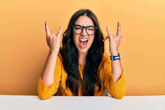 Beautiful brunette young woman wearing glasses and casual clothes sitting on the table shouting with crazy expression doing rock symbol with hands up. music star. heavy concept.