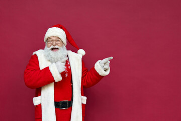 Funny happy excited old bearded Santa Claus face wearing costume looking at camera showing pointing fingers aside advertising Christmas promotion, New Year xmas discount ad isolated on red background. - obrazy, fototapety, plakaty