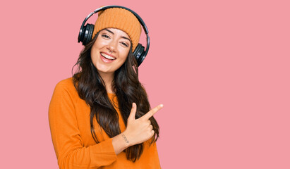 Beautiful brunette young woman listening to music using headphones cheerful with a smile of face pointing with hand and finger up to the side with happy and natural expression on face