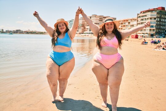 Two plus size overweight sisters twins women happy and proud of their bodies walking at the beach on summer holidays