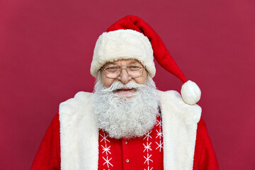 Close up face headshot portrait of funny happy old bearded Santa Claus face wearing hat, glasses, looking at camera, laughing, standing on Merry Christmas, Happy New Year isolated on red background.