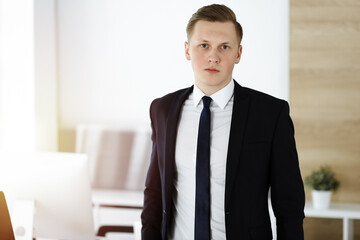 Cheerful businessman standing and looking at camera in sunny office. Headshot of young entrepreneur