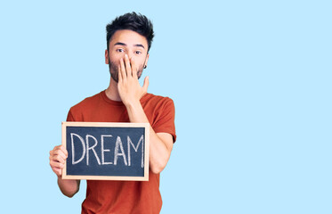 Young hispanic man holding blackboard with dream word covering mouth with hand, shocked and afraid...