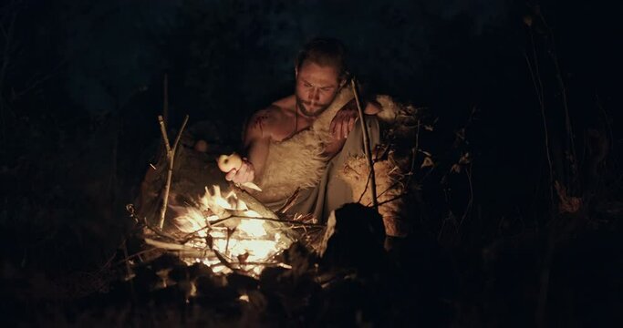 Primitive hunter gatherer wearing sheepskins seated on the ground eating by the light of a flaming fire at night lit by the glowing coals