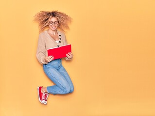 Young beautiful curly student girl wearing glasses smiling happy. Jumping with smile on face reading book over isolated yellow background.