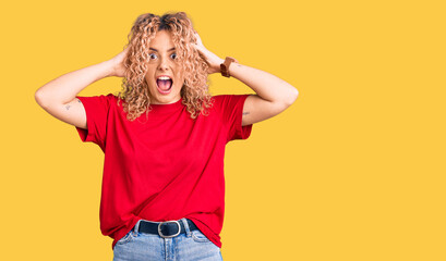 Young blonde woman with curly hair wearing casual red tshirt crazy and scared with hands on head,...