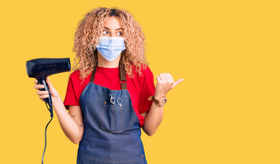 Young blonde woman with curly hair holding dryer blow wearing safety mask for coranvirus pointing thumb up to the side smiling happy with open mouth