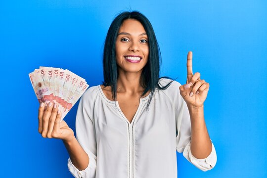 Young african american woman holding 10 colombian pesos banknotes with a big smile on face, pointing with hand finger to the side looking at the camera.