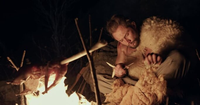 Primitive hunter gatherer cooking his meat on a campfire at night with a small animal carcass roasting on a spit over the flames as he sits sharpening a tool by hand - wild caveman