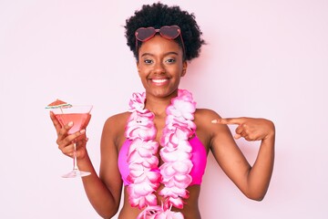 Young african american woman wearing bikini and hawaiian lei holding cocktail pointing finger to...