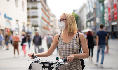 Woman walking by her bicycle on pedestrian city street wearing medical face mask in public to prevent spreading of corona virus. New normal during covid epidemic. Social responsibility.