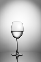 A wine glass with a transparent liquid stands on a mirror surface against the background of a light spot.