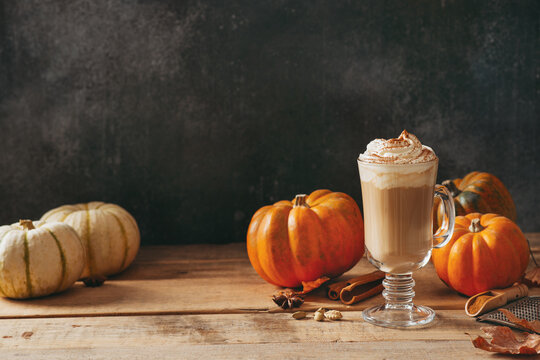 Spice pumpkin latte with cream foam, cinnamon stick, leaf and orange pumpkins on wooden background