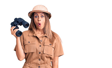 Young beautiful woman wearing explorer hat holding binoculars scared and amazed with open mouth for...