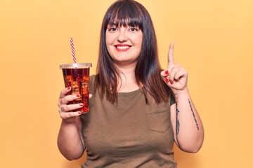 Young plus size woman drinking glass of cola beverage smiling with an idea or question pointing finger with happy face, number one