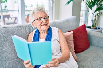 Fototapeta na wymiar Elder senior woman with grey hair smiling happy sitting on the sofa reading a book at home