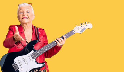 Senior beautiful woman with blue eyes and grey hair wearing a modern look playing electric guitar smiling friendly offering handshake as greeting and welcoming. successful business.