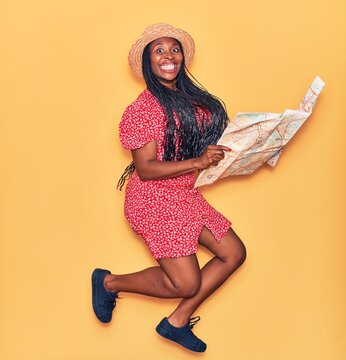 Young beautiful african american woman wearing summer clothes and hat smiling happy. Jumping with smile on face holding city map over isolated yellow background.