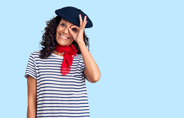 Middle age beautiful woman wearing french beret and scarf smiling happy doing ok sign with hand on eye looking through fingers