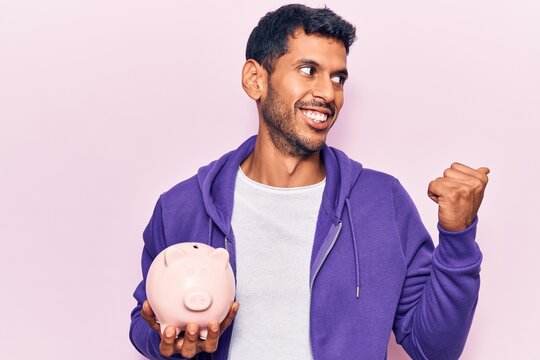 Young latin man holding piggy bank pointing thumb up to the side smiling happy with open mouth