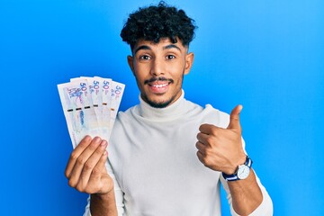 Young arab handsome man holding 50 colombian pesos banknotes smiling happy and positive, thumb up...