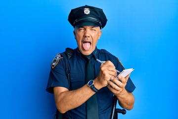 Handsome middle age mature man wearing police uniform writing a ticket sticking tongue out happy with funny expression.