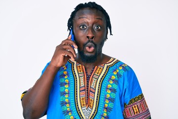 Young african american man with braids wearing ethnic tshirt having conversation talking on the smartphone scared and amazed with open mouth for surprise, disbelief face