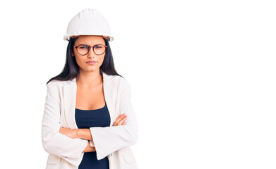 Young beautiful latin girl wearing architect hardhat and glasses skeptic and nervous, disapproving expression on face with crossed arms. negative person.
