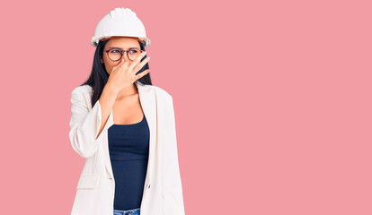 Young beautiful latin girl wearing architect hardhat and glasses smelling something stinky and...