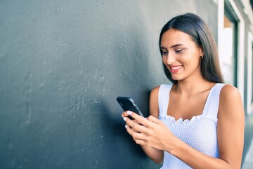 Young latin girl smiling happy using smartphone leaning on the wall.