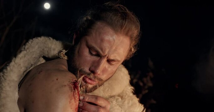 Primitive man clad in sheepskin scratching at a bleeding wound on his shoulder with a stick to use it as pigment to write or draw by the glowing light of a fire at night - wild caveman