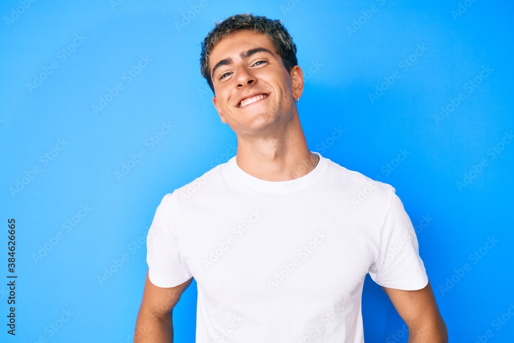 Wall mural young handsome man wearing casual white t shirt looking positive and happy standing and smiling with