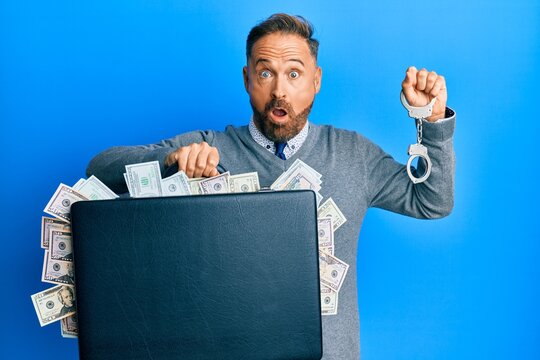 Handsome middle age man holding briefcase full of dollars and handcuffs afraid and shocked with surprise and amazed expression, fear and excited face.
