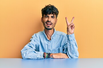 Young hispanic man wearing casual clothes sitting on the table smiling with happy face winking at the camera doing victory sign. number two.